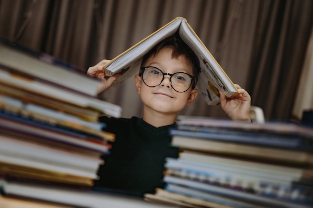 Ragazzo caucasico carino con gli occhiali che copre la testa con un libro Ragazzo allegro che sbircia da dietro pile di...