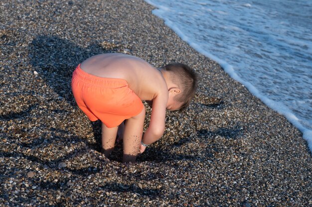 Ragazzo caucasico del bambino che gioca sulla spiaggia in una giornata di sole. Il bambino sta scavando una buca.