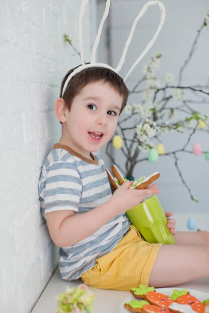 Ragazzo caucasico del bambino con le orecchie divertenti del coniglietto mangia i biscotti Ragazzo con i biscotti vicino all'albero di Pasqua su una priorità bassa bianca