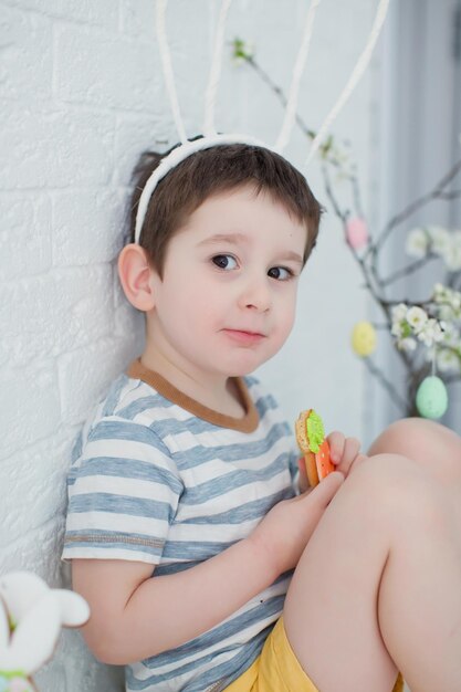 Ragazzo caucasico del bambino con le orecchie divertenti del coniglietto mangia i biscotti Ragazzo con i biscotti vicino all'albero di Pasqua su una priorità bassa bianca