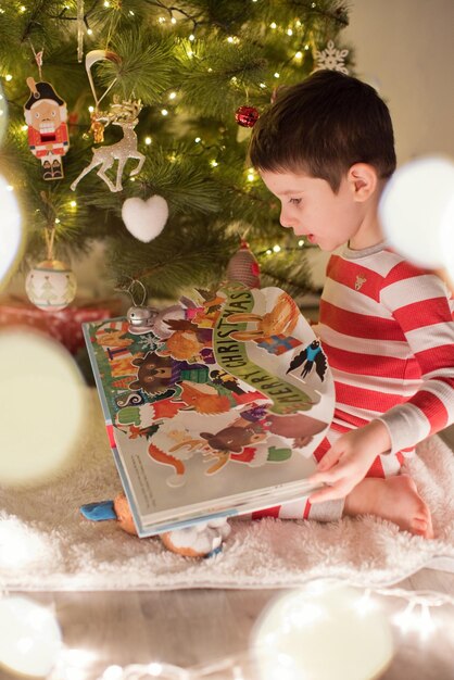 Ragazzo caucasico in pigiama a righe con un libro seduto vicino all'albero di Natale Interiore accogliente di Natale con decorazioni e luci