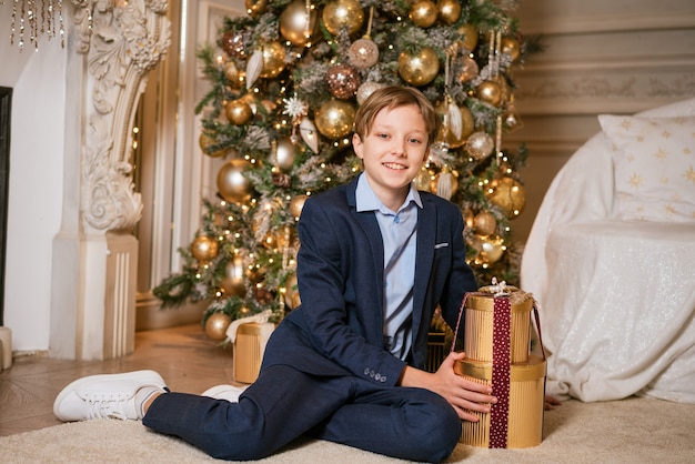 Ragazzo caucasico in tuta si siede vicino al regalo di natale adolescente carino accanto al regalo per natale aspetta fino a...