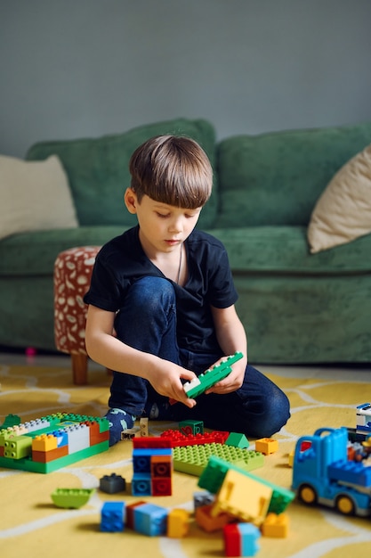 Ragazzo caucasico prescolare che gioca con il costruttore seduto sul pavimento, un sacco di costruttore di blocchi di plastica colorati