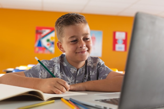 Ragazzo caucasico seduto a una scrivania in aula scrivendo, guardando il laptop e sorridendo