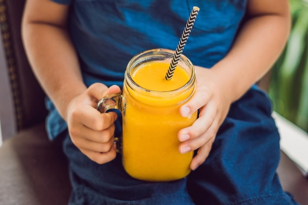 Ragazzo che beve frullato succoso dal mango in barattolo di vetro con paglia rossa a strisce su fondo di legno vecchio. Concetto di vita sana, copia spazio