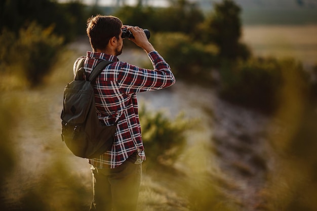 Ragazzo che guarda il binocolo in collina uomo in maglietta con zaino