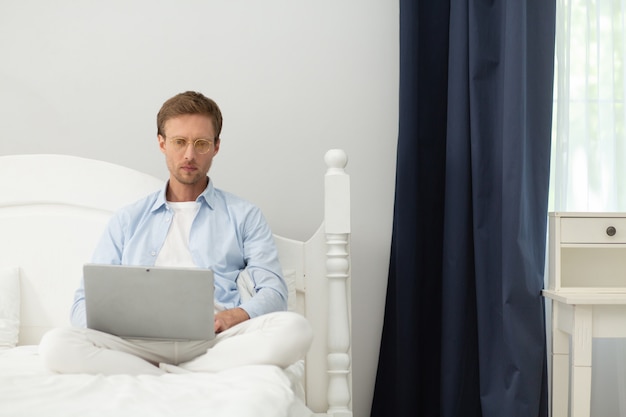 Ragazzo che lavora con il computer portatile nel suo letto a casa. Pigiama calmo e tranquillo di mezza età uomo che indossa pigiama e sottili occhiali trasparenti seduto nel suo letto sotto la coperta e lavorando con il computer