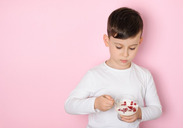 Ragazzo che mangia yogurt da dessert con ribes in una maglietta bianca su una superficie di studio rosa