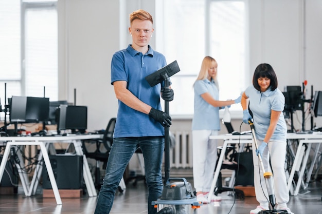 Ragazzo con aspirapolvere Gruppo di lavoratori puliscono insieme l'ufficio moderno durante il giorno