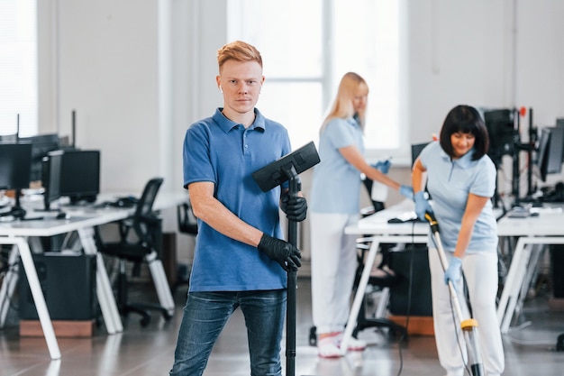 Ragazzo con aspirapolvere Gruppo di lavoratori puliscono insieme l'ufficio moderno durante il giorno