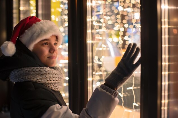 Ragazzo con cappello di Babbo Natale che guarda e sogna nella vetrina illuminata del negozio