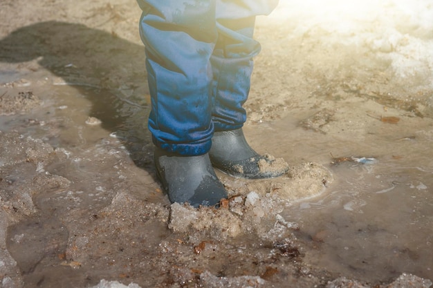 Ragazzo con i piedi in stivali di gomma nella pozzanghera fangosa con sabbia e neve Bambino ragazzo di 67 anni che cammina e gioca con stivali di gomma in terra fusa La primavera è arrivata