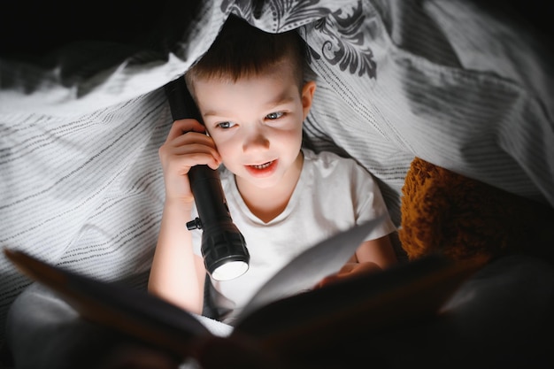 Ragazzo con il libro di lettura della torcia elettrica sotto la coperta a casa