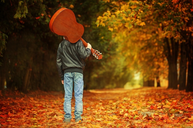 Ragazzo con la chitarra che cammina sulla strada di autunno. Vista posteriore