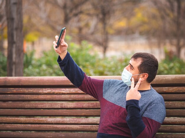 Ragazzo con maschera protettiva che si fa un selfie su una panchina del parco al tramonto