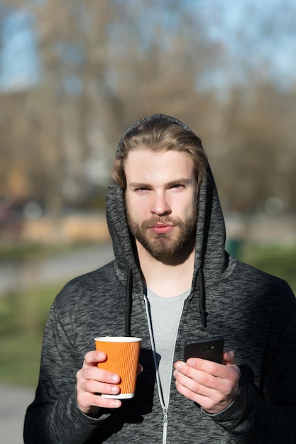 Ragazzo con smartphone e bevanda da asporto all'aria aperta. L'uomo barbuto nel cappuccio tiene la tazza di caffè usa e getta all'aperto soleggiato. Umore del caffè o del tè. Tè del mattino a colazione. Nuova tecnologia e vita moderna.