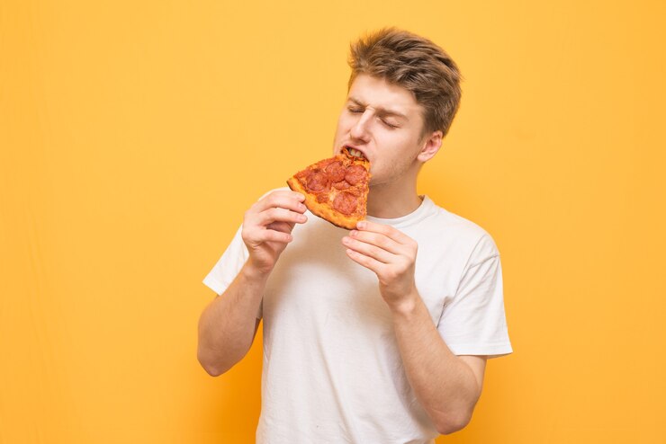 Ragazzo con una maglietta bianca mangia un appetitoso pezzo di pizza con gli occhi chiusi su un giallo
