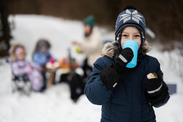 Ragazzo con una tazza di tè a portata di mano contro la sua famiglia nella foresta invernale che trascorre del tempo insieme in un picnic