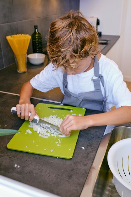 Ragazzo concentrato in grembiule che taglia cipolla cruda con un coltello mentre cucina a tavola in casa