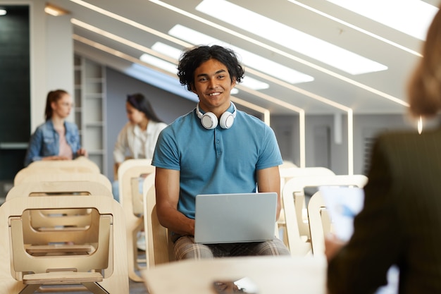 Ragazzo curioso sorridente della corsa mista con le cuffie senza fili intorno al collo che si siede con il computer portatile sulla sedia e parlando con l'insegnante universitario