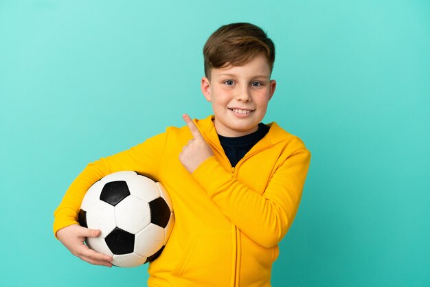 Ragazzo dai capelli rossi che gioca a calcio isolato su sfondo blu che punta al lato per presentare un prodotto