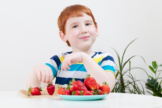 Ragazzo dai capelli rossi che mangia fragole rosse mature, primo piano bambino felice