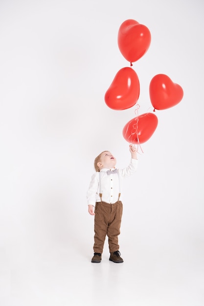 Ragazzo del bambino del bambino in vestito che tiene palloncino cuore su bianco.