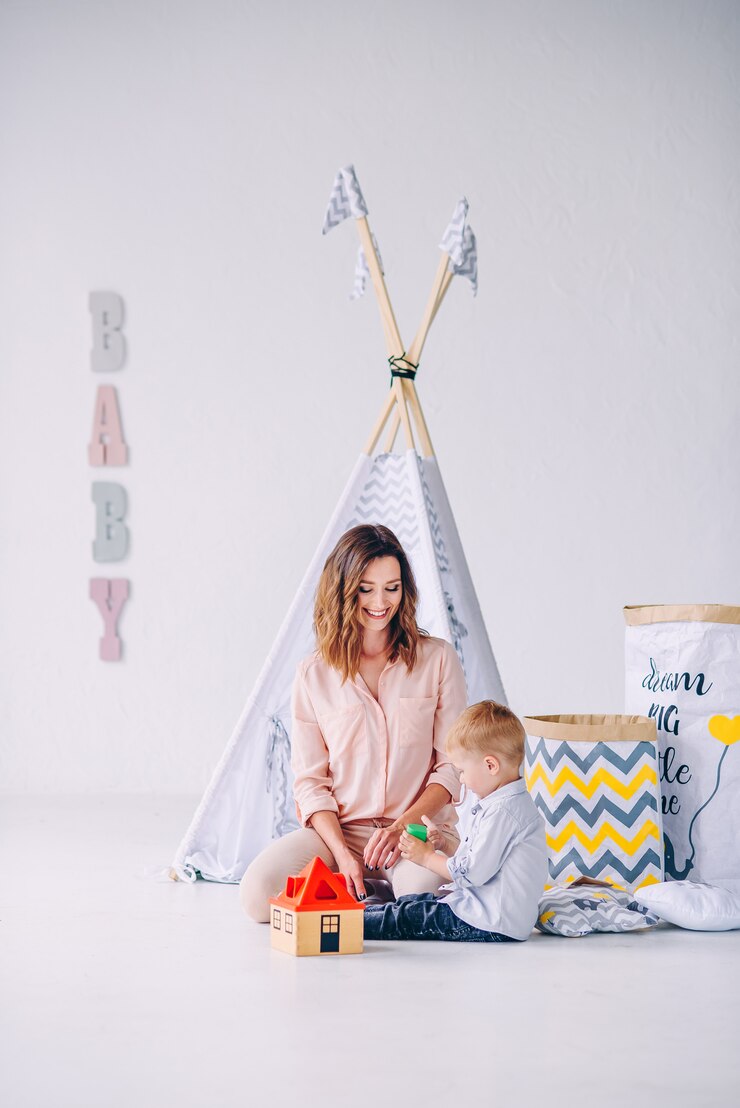 Ragazzo del bambino e sua bella giovane madre che giocano insieme ai giocattoli educativi nella stanza leggera con il wigwam dei bambini e le eco-borse per i giocattoli.