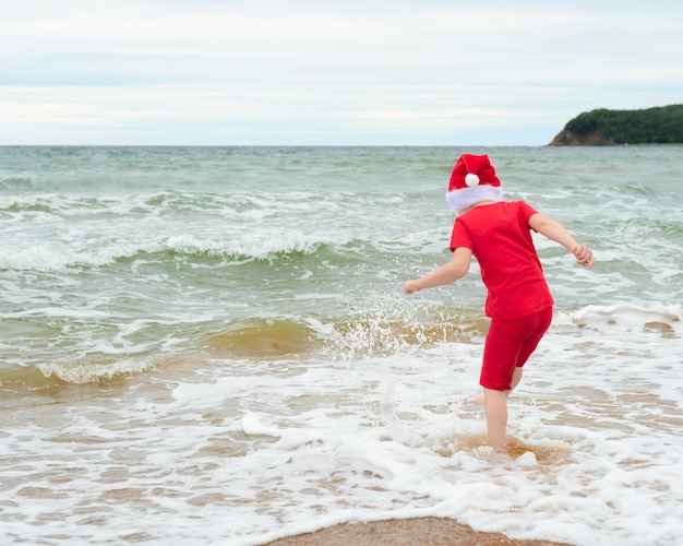 Ragazzo del bambino in panno rosso e cappello di Babbo Natale che si gode il Natale sulla spiaggia durante le vacanze di Natale concetto di celebrazione di Natale o Capodanno