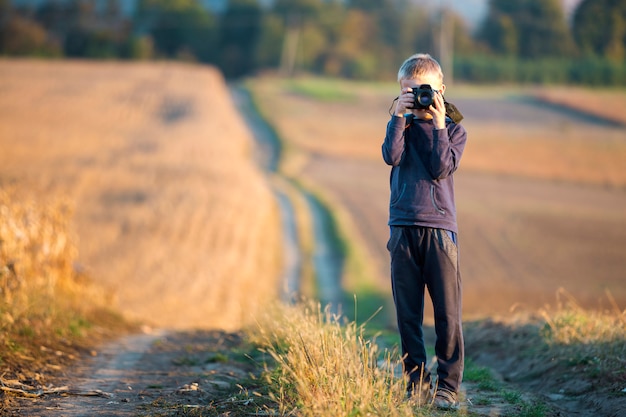 Ragazzo del bambino piccolo con la macchina fotografica della foto che prende immagine del giacimento di grano su rurale vago.