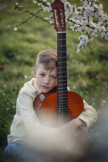 Ragazzo del ritratto con la chitarra che si siede sull&#39;erba nel giorno di estate.