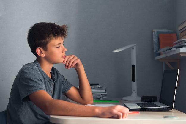 Ragazzo dell'adolescente dello studente che fa i compiti con il quaderno aperto del computer portatile e il posto di lavoro del computer a casa