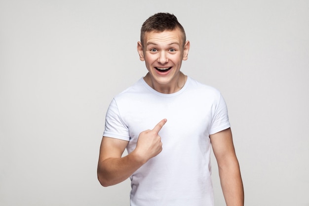 Ragazzo di felicità che guarda l'obbiettivo e che punta il dito se stesso. Foto in studio, muro grigio