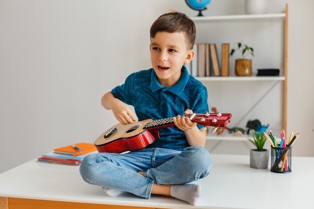 Ragazzo di talento che suona l'ukulele soprano seduto sulla scrivania Ragazzo in età prescolare che impara la chitarra a piacimento