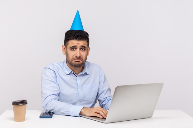 Ragazzo dipendente infelice con cono divertente sulla testa seduto nel posto di lavoro dell'ufficio utilizzando il laptop e guardando depresso sconvolto per il fine settimana annullato studio indoor girato isolato su sfondo bianco