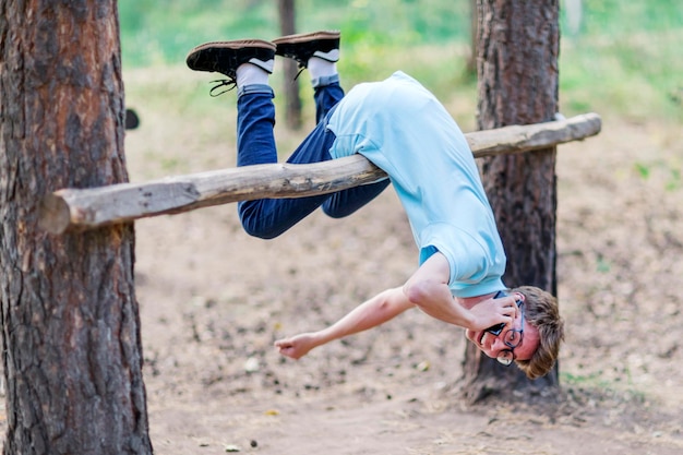 Ragazzo divertente appeso a testa in giù su una trave nella foresta e parlando al telefono