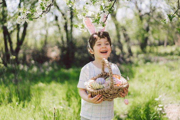Ragazzo divertente con cesto di uova e orecchie da coniglio a caccia di uova di Pasqua nel soleggiato giardino primaverile
