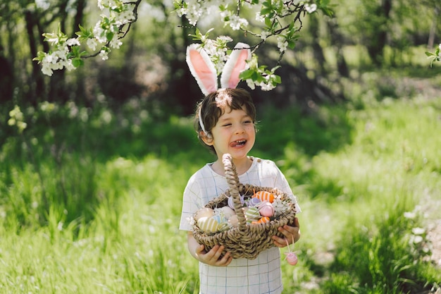 Ragazzo divertente con cesto di uova e orecchie da coniglio durante la caccia alle uova di Pasqua nel soleggiato giardino primaverile