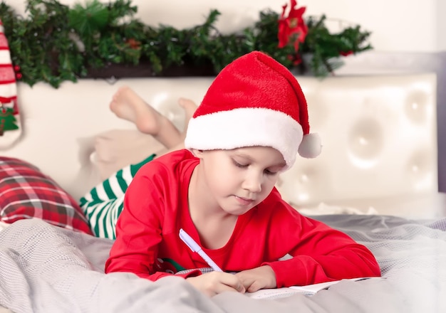 Ragazzo divertente con un cappello da Babbo Natale che scrive una lettera a Babbo Natale sdraiato su un letto