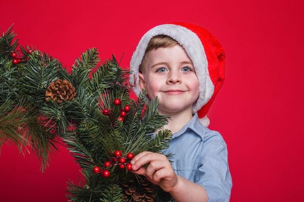 Ragazzo divertente in cappello di Babbo Natale con ornamento di Natale su sfondo rosso.