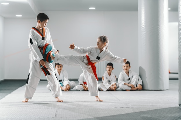 Ragazzo e ragazza caucasici in dobok che hanno addestramento di taekwondo in palestra. Ragazza che dà dei calci mentre ragazzo che tiene il bersaglio di scossa.