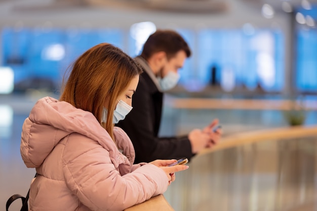 Ragazzo e ragazza in maschera utilizzano il telefono cellulare nel centro commerciale della città in tempo di pandemia