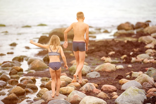 Ragazzo e ragazza sul litorale di pietra Fratello e la sua sorellina vista posteriore posteriore bambini che giocano a catturare sulla riva del mare rocce ricoperte di alghe alghe Bambini sullo sfondo dell'orizzonte del mare