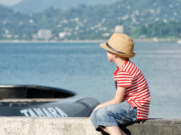 Ragazzo è seduto sulla spiaggia con un cappello e una maglietta a strisce, guardando la nave.