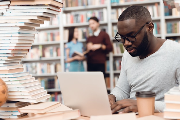 Ragazzo etnico afroamericano circondato da libri in biblioteca. Lo studente usa il laptop e beve il caffè.