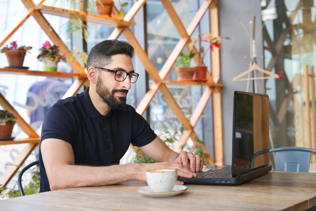 Ragazzo etnico musulmano arabo giovane freelance seduto nella caffetteria che lavora al computer portatile bere caffè nella caffetteria