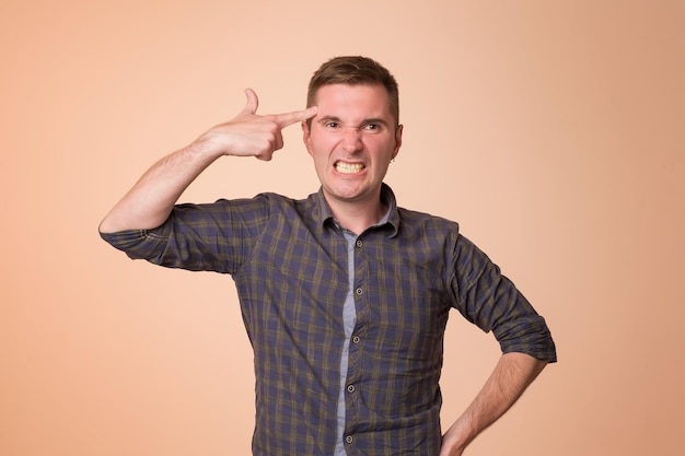 Ragazzo europeo stanco o annoiato che tiene il gesto della pistola vicino alle tempie e fa le facce mentre si trova su sfondo arancione