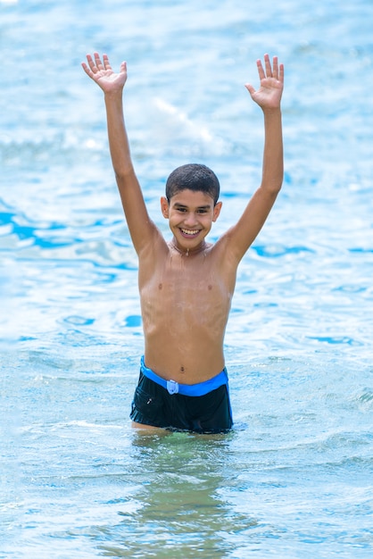 Ragazzo felice attivo che gioca in acqua di mare blu con le braccia in su