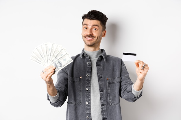 Ragazzo felice che mostra carta di credito in plastica e banconote da un dollaro, dando scelta, in piedi su sfondo bianco.