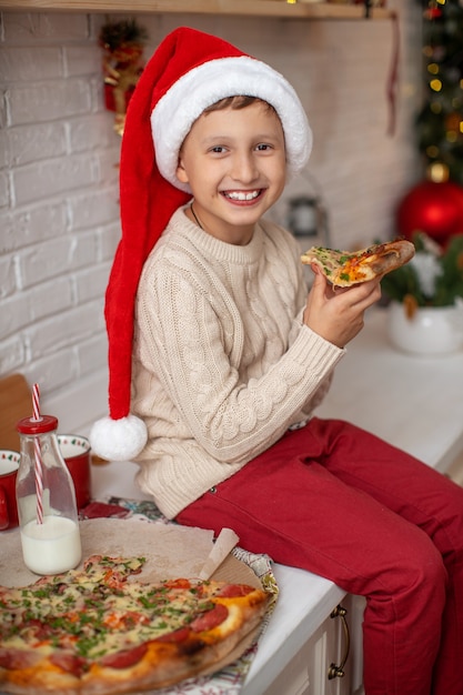 Ragazzo felice con il cappello della santa che mangia pizza in cucina